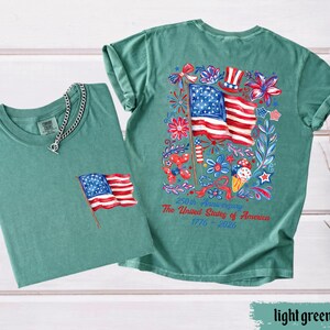 May include: Two light green t-shirts. One shirt has a small American flag embroidered on the pocket. The other shirt has a large graphic of the American flag with floral and patriotic elements. Text on the back reads "250th Anniversary The United States of America 1776-2026."