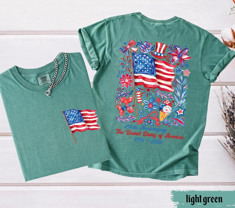 May include: Two light green t-shirts. One shirt has a small American flag embroidered on the pocket. The other shirt has a large graphic of the American flag with floral and patriotic elements. Text on the back reads "250th Anniversary The United States of America 1776-2026."