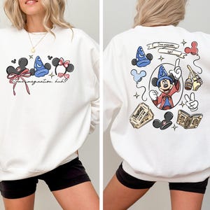 May include: White sweatshirt featuring Disney-themed graphics. The front displays Mickey Mouse ears, a Sorcerer's hat, and Minnie Mouse ears with text that reads "Some imagination, huh?". The back showcases Mickey Mouse as a sorcerer, along with other Disney icons.