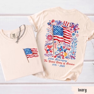 May include: Ivory-colored t-shirts with American flag designs. One shirt has a small flag on the pocket, the other features a large flag with floral and celebratory graphics. Text on the back reads "250th Anniversary The United States of America 1776-2026."