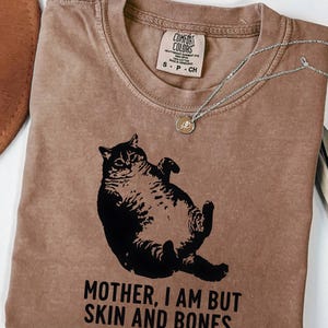 May include: Espresso-colored t-shirt featuring a black graphic of a cat lying on its back. The text "MOTHER, I AM BUT SKIN AND BONES" is printed below the cat. A silver necklace with a small pendant rests on the shirt.