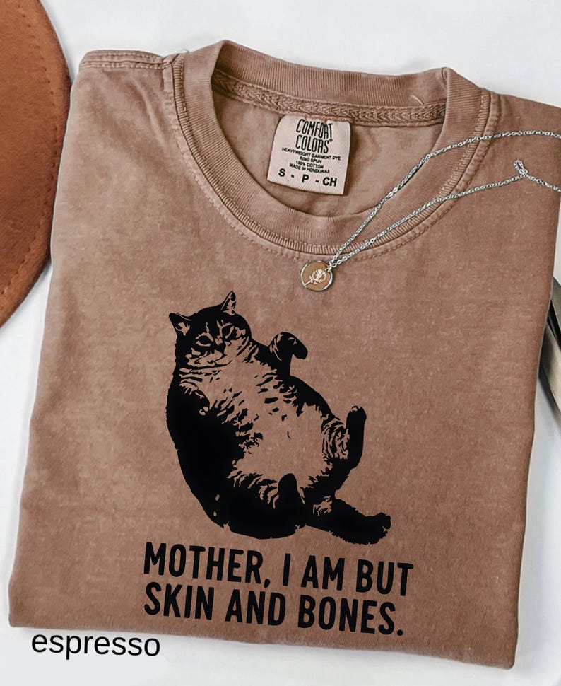 May include: Espresso-colored t-shirt featuring a black graphic of a cat lying on its back. The text "MOTHER, I AM BUT SKIN AND BONES" is printed below the cat. A silver necklace with a small pendant rests on the shirt.