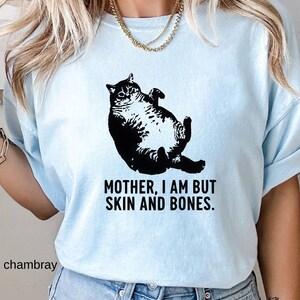 May include: Light blue t-shirt featuring a black illustration of a cat lying on its back. The text "MOTHER, I AM BUT SKIN AND BONES" is printed below the cat. The word "chambray" is printed on the bottom left.