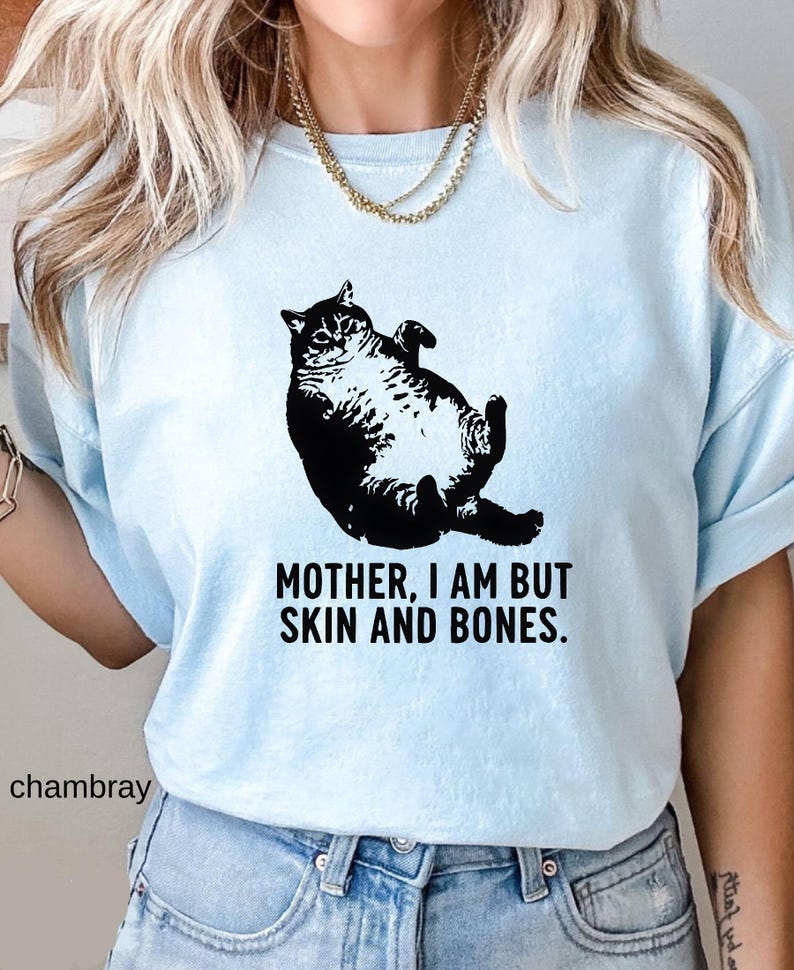 May include: Light blue t-shirt featuring a black illustration of a cat lying on its back. The text "MOTHER, I AM BUT SKIN AND BONES" is printed below the cat. The word "chambray" is printed on the bottom left.