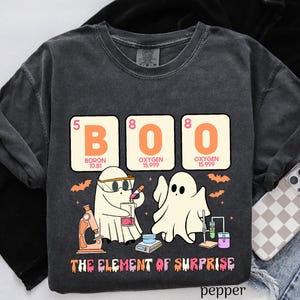 May include: Dark gray t-shirt with a Halloween-themed graphic. The design features the word "BOO" spelled out with periodic table elements, along with cartoon ghosts and the phrase "THE ELEMENT OF SURPRISE".