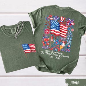 May include: Two moss green t-shirts. One has a small American flag patch, the other has a large colorful graphic of the American flag, flowers, and text that reads "250th Anniversary The United States of America 1776-2026".