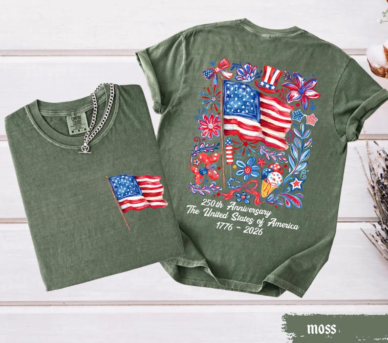 May include: Two moss green t-shirts. One has a small American flag patch, the other has a large colorful graphic of the American flag, flowers, and text that reads "250th Anniversary The United States of America 1776-2026".