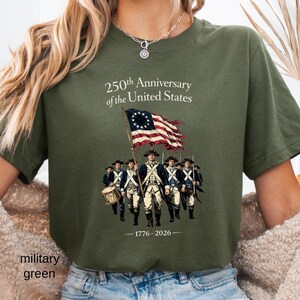 May include: Military green t-shirt featuring a graphic commemorating the 250th anniversary of the United States. The design includes an illustration of soldiers, a flag, and the years 1776-2026.