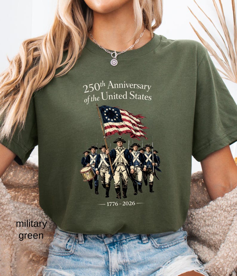 May include: Military green t-shirt featuring a graphic commemorating the 250th anniversary of the United States. The design includes an illustration of soldiers, a flag, and the years 1776-2026.