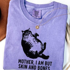 May include: A violet Comfort Colors t-shirt featuring a black graphic of a cat lying on its back. The text "MOTHER, I AM BUT SKIN AND BONES" is printed below the cat. A gold necklace with a pendant is draped across the shirt.