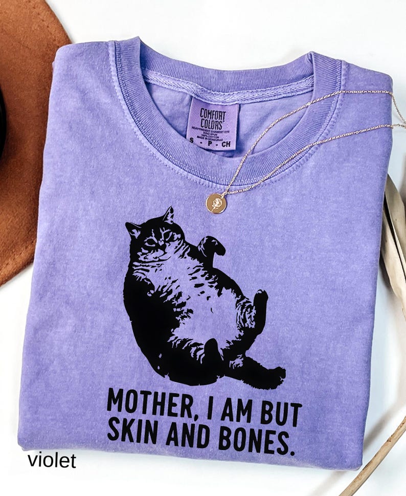 May include: A violet Comfort Colors t-shirt featuring a black graphic of a cat lying on its back. The text "MOTHER, I AM BUT SKIN AND BONES" is printed below the cat. A gold necklace with a pendant is draped across the shirt.