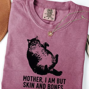 May include: Berry-colored t-shirt with a black graphic of a cat lying on its back. The shirt has the text "MOTHER, I AM BUT SKIN AND BONES." A gold-colored necklace with a pendant is draped across the shirt.