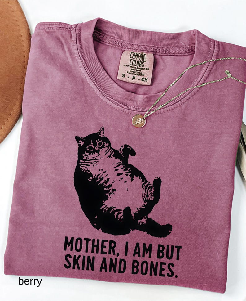 May include: Berry-colored t-shirt with a black graphic of a cat lying on its back. The shirt has the text "MOTHER, I AM BUT SKIN AND BONES." A gold-colored necklace with a pendant is draped across the shirt.