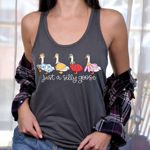 K&ouml;nnte beinhalten: Dunkelgraues Tanktop mit einem skurrilen Design von vier G&auml;nsen, jede mit einem einzigartigen Outfit, darunter ein Blumentopf, ein gelbes Cape, eine Erdbeere und ein rosa Rock. Der Text "just a silly goose" steht darunter.