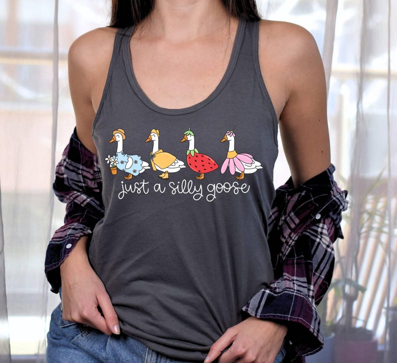 K&ouml;nnte beinhalten: Dunkelgraues Tanktop mit einem skurrilen Design von vier G&auml;nsen, jede mit einem einzigartigen Outfit, darunter ein Blumentopf, ein gelbes Cape, eine Erdbeere und ein rosa Rock. Der Text "just a silly goose" steht darunter.