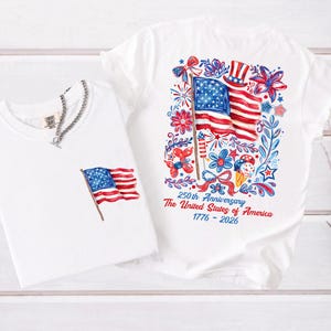 May include: Two white t-shirts with patriotic designs. One shirt features a small American flag graphic, and the other displays a larger flag design with floral and firework elements. Text on the shirt reads "250th Anniversary The United States of America 1776-2026."