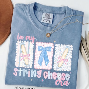 May include: Blue jean Comfort Colors t-shirt with the phrase "in my string cheese cra" and illustrations of string cheese and bows. A gold necklace with a small pendant is draped across the shirt.