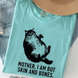 May include: A light blue t-shirt with a black graphic of a cat lying on its back. The text "MOTHER, I AM BUT SKIN AND BONES" is printed below the cat. The shirt is labeled "chalky mint".