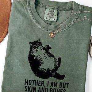 May include: A moss green t-shirt with a black graphic of a cat lying on its back. The text "MOTHER, I AM BUT SKIN AND BONES" is printed below the cat. A silver necklace with a small pendant is draped across the shirt.