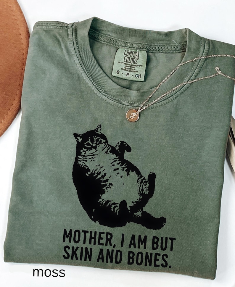 May include: A moss green t-shirt with a black graphic of a cat lying on its back. The text "MOTHER, I AM BUT SKIN AND BONES" is printed below the cat. A silver necklace with a small pendant is draped across the shirt.