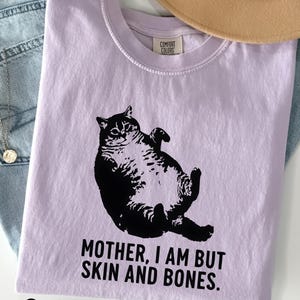 May include: A lilac-colored t-shirt with a black graphic of a cat lying on its back. The text "MOTHER, I AM BUT SKIN AND BONES" is printed below the cat. The word "Orchid" is printed at the bottom of the shirt.