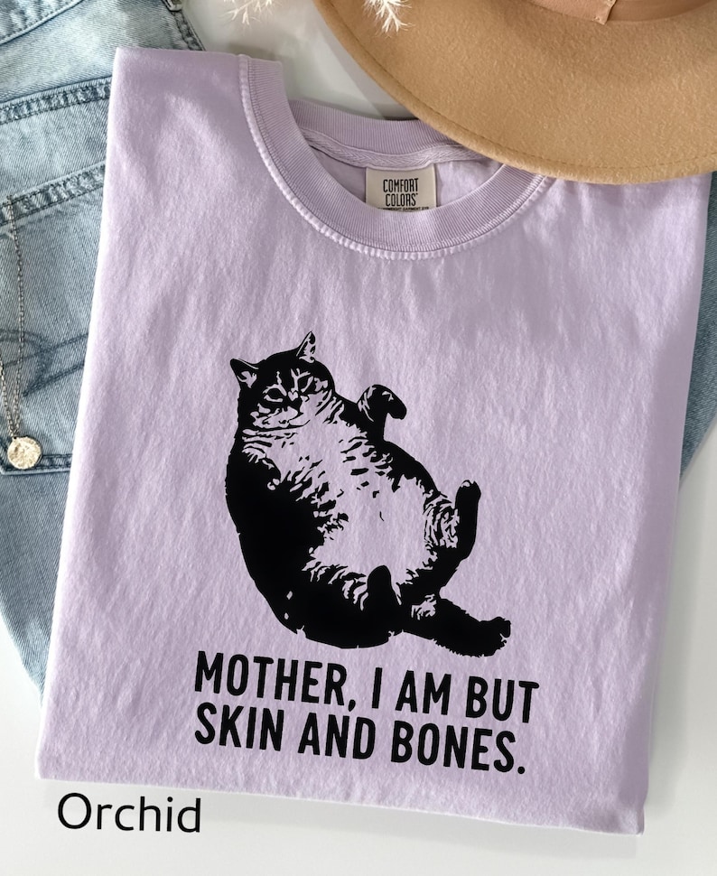 May include: A lilac-colored t-shirt with a black graphic of a cat lying on its back. The text "MOTHER, I AM BUT SKIN AND BONES" is printed below the cat. The word "Orchid" is printed at the bottom of the shirt.