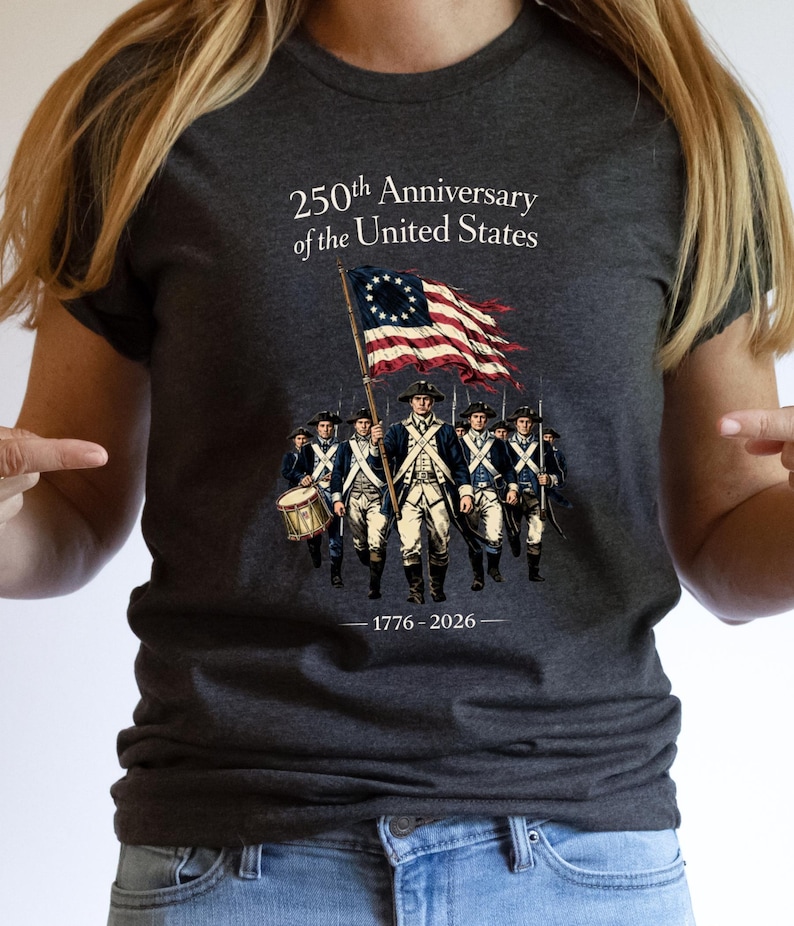 May include: Dark gray t-shirt featuring a graphic commemorating the 250th Anniversary of the United States. The design includes a colonial flag and a line of soldiers. The text "250th Anniversary of the United States" and "1776-2026" are also displayed.