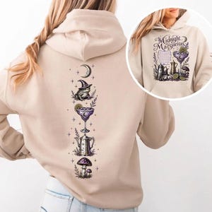 May include: A beige hooded sweatshirt featuring a vertical graphic print on the back and a matching chest print. The design includes a crescent moon, a toad wearing a witch hat, a margarita glass, a teapot, and a mushroom in purple and black ink.