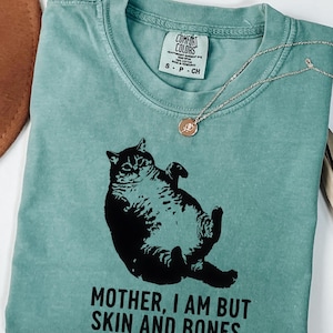 May include: Light green Comfort Colors t-shirt with a black graphic of a cat and the text "MOTHER, I AM BUT SKIN AND BONES." A silver necklace with a rose pendant is draped on the shirt. The shirt has a crew neck and short sleeves.