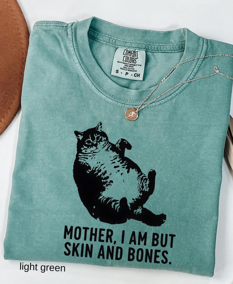 May include: Light green Comfort Colors t-shirt with a black graphic of a cat and the text "MOTHER, I AM BUT SKIN AND BONES." A silver necklace with a rose pendant is draped on the shirt. The shirt has a crew neck and short sleeves.