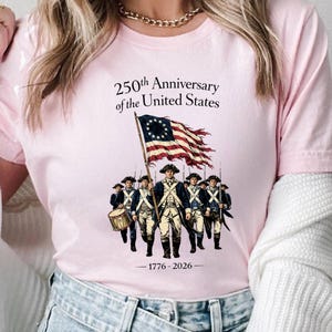 May include: Pink t-shirt featuring a graphic commemorating the 250th Anniversary of the United States. The design includes a colonial flag, soldiers, and the years 1776-2026. The shirt is made of a soft material.