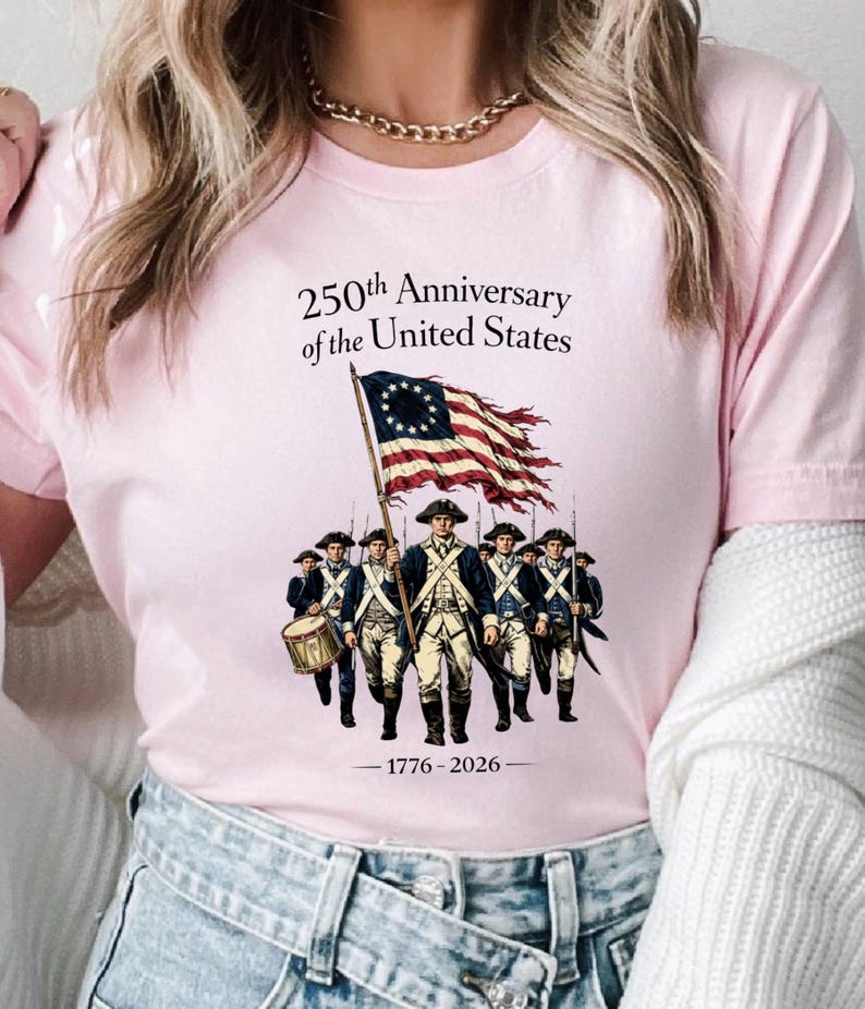 May include: Pink t-shirt featuring a graphic commemorating the 250th Anniversary of the United States. The design includes a colonial flag, soldiers, and the years 1776-2026. The shirt is made of a soft material.
