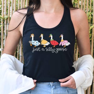 K&ouml;nnte beinhalten: Schwarzes Tanktop mit einer skurrilen Illustration von vier G&auml;nsen in verschiedenen Outfits, darunter ein Blumenkleid, ein gelber Mantel, ein Erdbeerkost&uuml;m und ein rosa Rock. Der Text "just a silly goose" ist darunter in Schreibschrift geschrieben.
