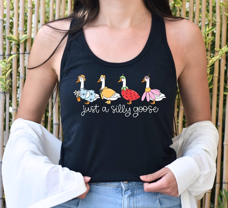 K&ouml;nnte beinhalten: Schwarzes Tanktop mit einer skurrilen Illustration von vier G&auml;nsen in verschiedenen Outfits, darunter ein Blumenkleid, ein gelber Mantel, ein Erdbeerkost&uuml;m und ein rosa Rock. Der Text "just a silly goose" ist darunter in Schreibschrift geschrieben.