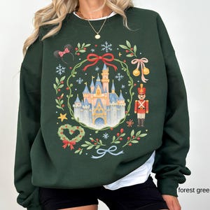 May include: Forest green sweatshirt featuring a festive design. The graphic includes a castle, snowflakes, a nutcracker, and a Minnie Mouse ear headband. A wreath of leaves and berries surrounds the castle. The text "forest green" is at the bottom right.