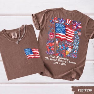 May include: Two espresso-colored t-shirts. One has a small American flag on the pocket. The other has a large graphic of the American flag with floral and firework designs. Text on the back reads "250th Anniversary The United States of America 1776-2026."