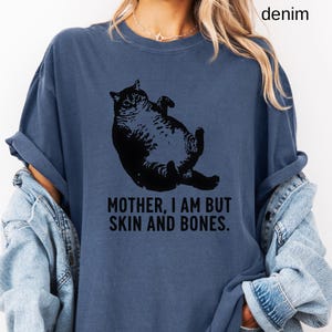 May include: Denim blue t-shirt featuring a black graphic of a cat lying on its back. The text "MOTHER, I AM BUT SKIN AND BONES" is printed below the cat. The shirt has a relaxed fit.