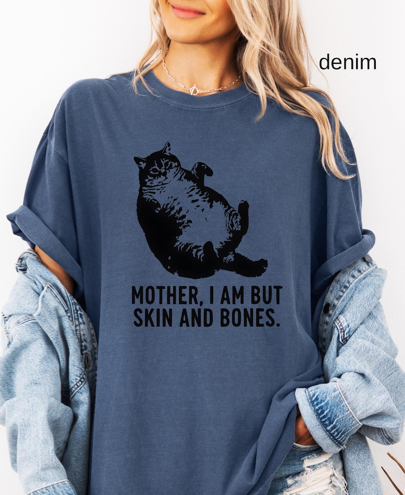 May include: Denim blue t-shirt featuring a black graphic of a cat lying on its back. The text "MOTHER, I AM BUT SKIN AND BONES" is printed below the cat. The shirt has a relaxed fit.