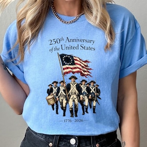 May include: Light blue t-shirt featuring a graphic commemorating the 250th anniversary of the United States. The design includes an illustration of soldiers, a waving American flag, and the years 1776-2026.