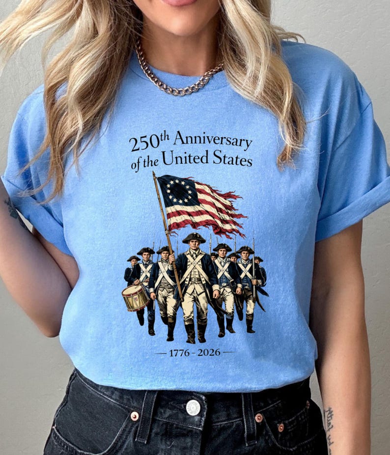May include: Light blue t-shirt featuring a graphic commemorating the 250th anniversary of the United States. The design includes an illustration of soldiers, a waving American flag, and the years 1776-2026.
