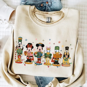 May include: A beige sweatshirt featuring a colorful graphic of Disney characters dressed as nutcrackers. The characters include Goofy, Minnie Mouse, Mickey Mouse, Daisy Duck, Donald Duck, and Pluto. The design includes snowflakes and Christmas lights.