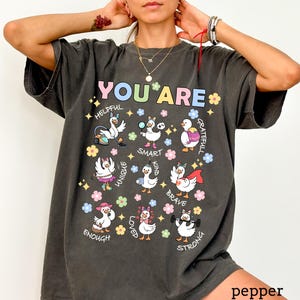 May include: Dark gray t-shirt with the words "YOU ARE" in colorful letters. The shirt features cartoon geese with positive affirmations like "helpful," "smart," and "brave." The shirt is a comfortable, casual style.
