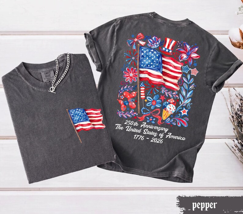 May include: Two dark gray t-shirts with American flag designs. One shirt has a small flag on the pocket, and the other has a large flag design on the back with the text "250th Anniversary The United States of America 1776-2026".
