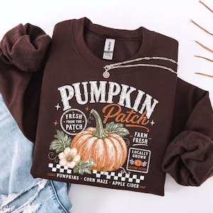 May include: A dark brown crewneck sweatshirt featuring a graphic print of an orange pumpkin with green leaves and text that reads PUMPKIN Patch, FRESH FROM THE PATCH, FARM FRESH, LOCALLY GROWN, AUTUMN PICKED, PUMPKINS, CORN MAZE, and APPLE CIDER.