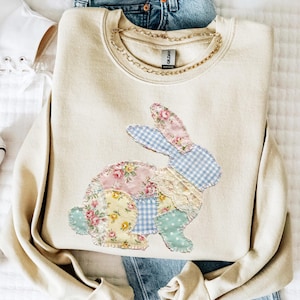 May include: A beige sweatshirt featuring a patchwork bunny design. The bunny is composed of floral, gingham, and lace fabrics in pastel colors. A gold chain necklace is around the neckline. The sweatshirt is folded on top of blue jeans.