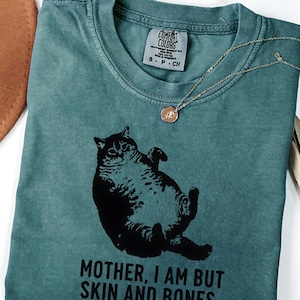 May include: Blue spruce t-shirt with a black graphic of a cat and the text "MOTHER, I AM BUT SKIN AND BONES." A gold-colored necklace with a pendant is draped on the shirt. The shirt has a Comfort Colors tag.