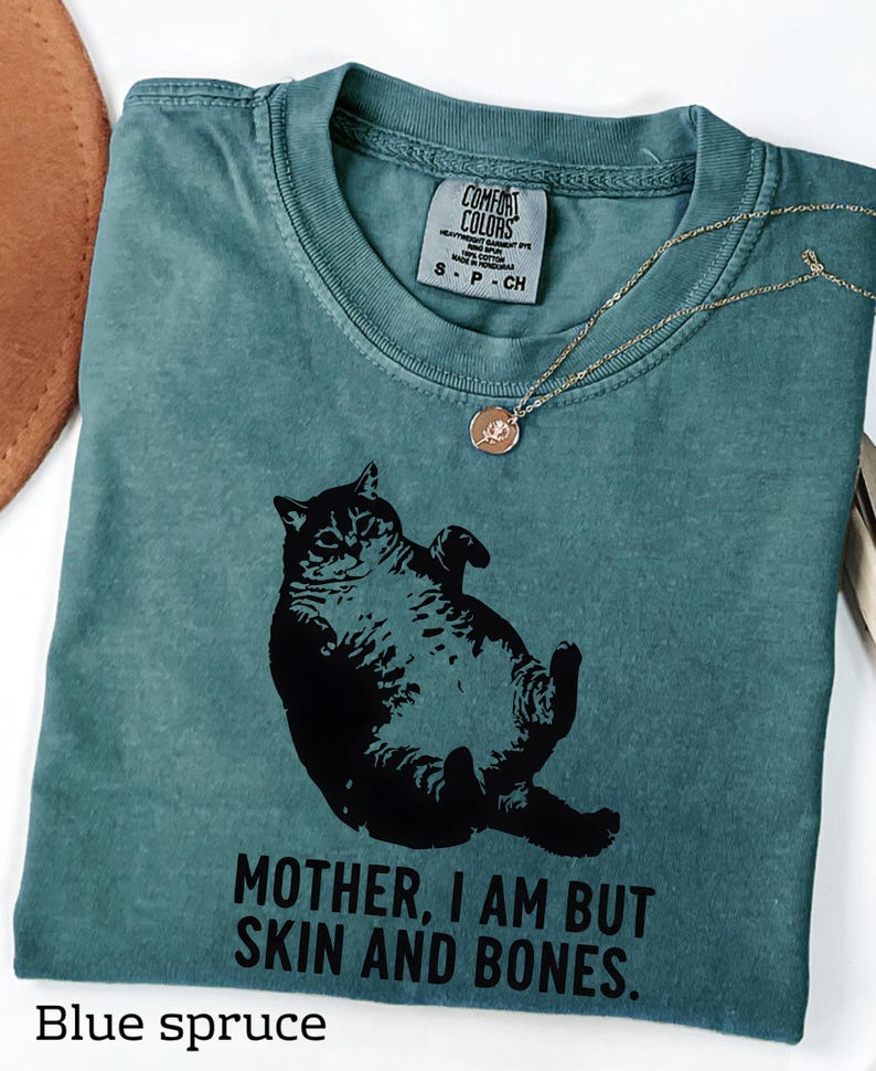 May include: Blue spruce t-shirt with a black graphic of a cat and the text "MOTHER, I AM BUT SKIN AND BONES." A gold-colored necklace with a pendant is draped on the shirt. The shirt has a Comfort Colors tag.