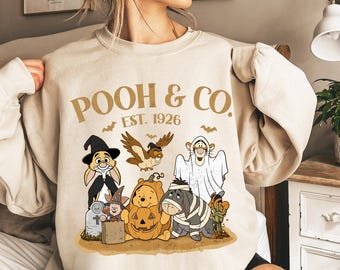 May include: Beige sweatshirt featuring a Halloween-themed graphic of Winnie the Pooh and friends. The design includes characters dressed in costumes, the text "POOH & CO. EST. 1926", and bat illustrations.