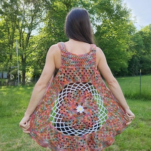May include: A colorful, handmade crochet vest with a circular white and floral design on the back. The vest features a mix of orange, red, green, and blue yarn. The sleeveless top is worn outdoors in a natural setting.