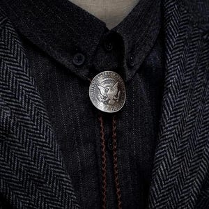 K&ouml;nnte beinhalten: Eine silberne Bolo-Krawatte mit einem Design einer Half-Dollar-M&uuml;nze der Vereinigten Staaten von Amerika. Die M&uuml;nze ist an einer braunen geflochtenen Kordel befestigt, vor einem dunkelgrauen Hemd und einer Jacke mit Fischgr&auml;tenmuster. Ein klassisches Accessoire.
